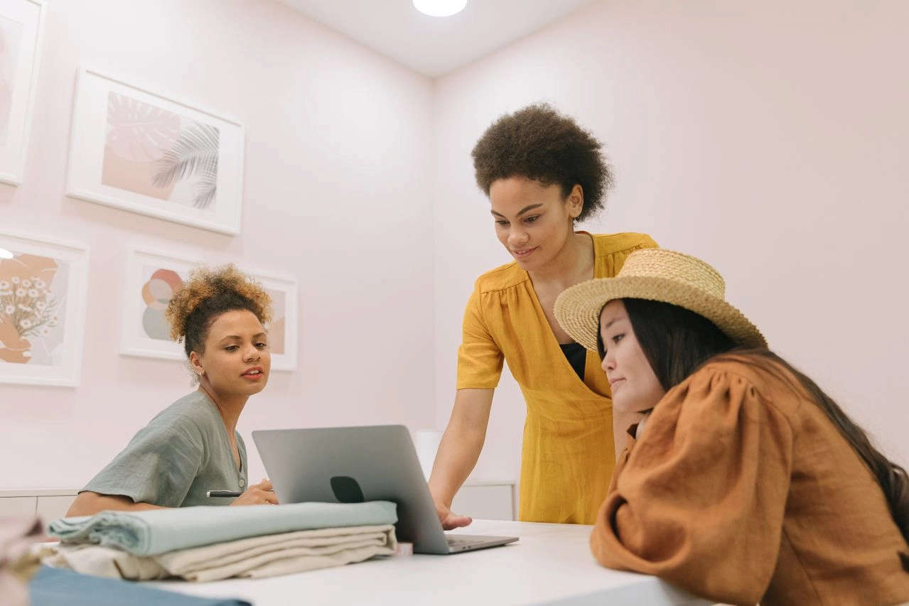 Three diverse women collaborating in a modern office setting with a laptop, showcasing teamwork. Business grants for women in Arkansas